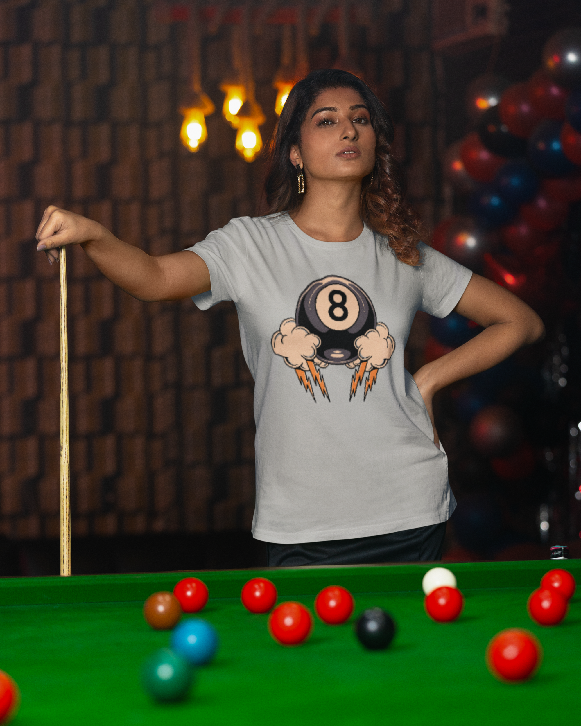 Woman in a gray t-shirt with a pool ball design standing behind a pool table.