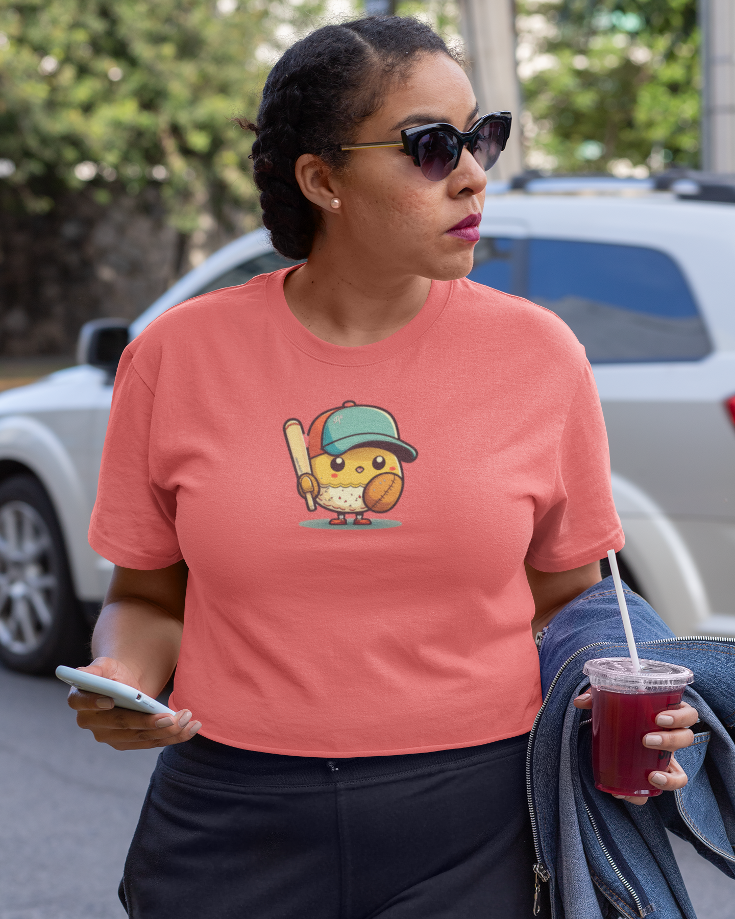 Woman wearing a pink t-shirt with a graphic design, holding a drink and phone on a street.
