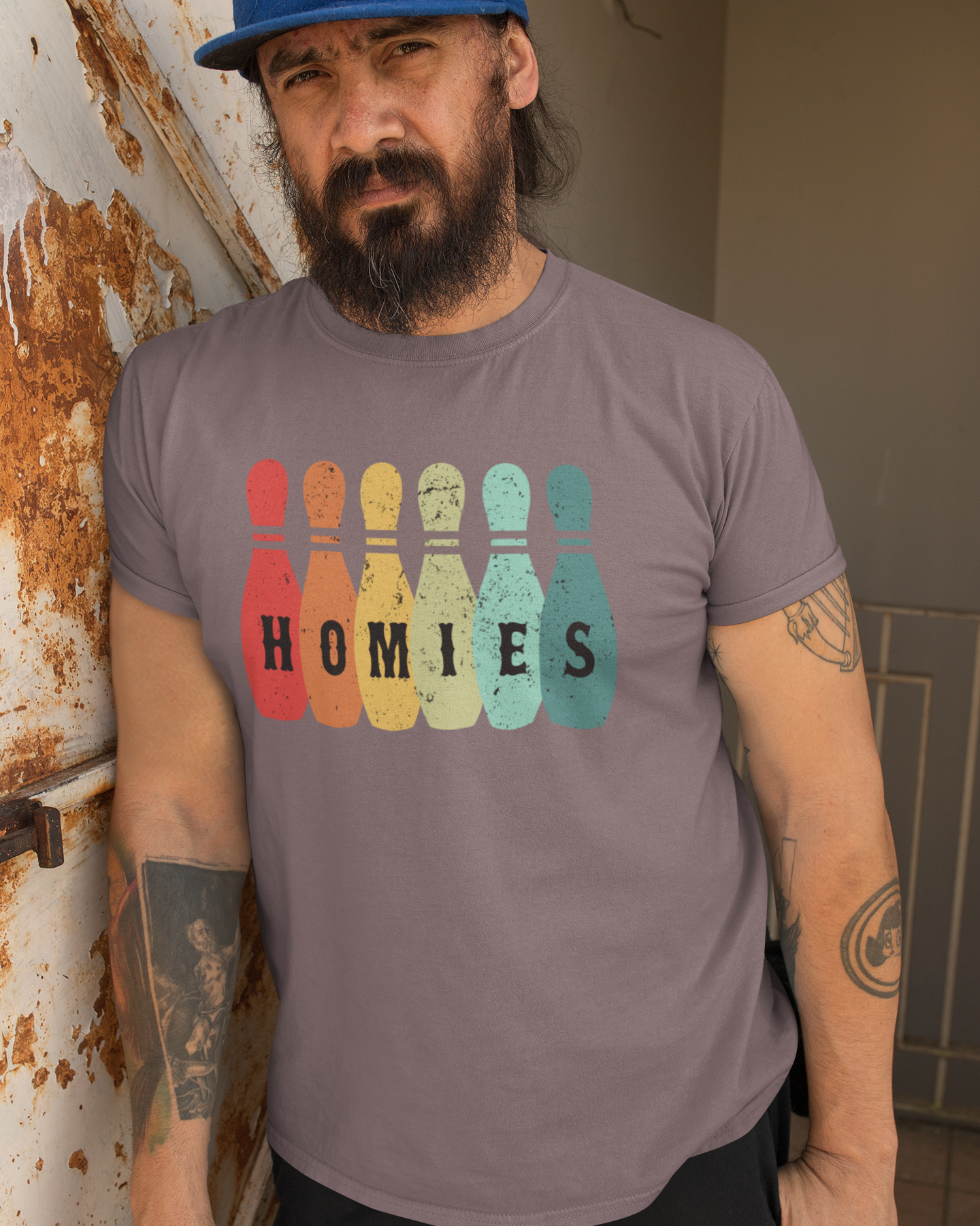 Man wearing a gray t-shirt with 'HOMIES' graphic design leaning against a wall.