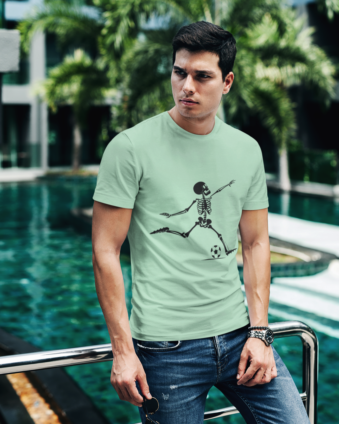 Man wearing a light green t-shirt with a skeleton playing soccer design, standing by a pool with greenery in the background.