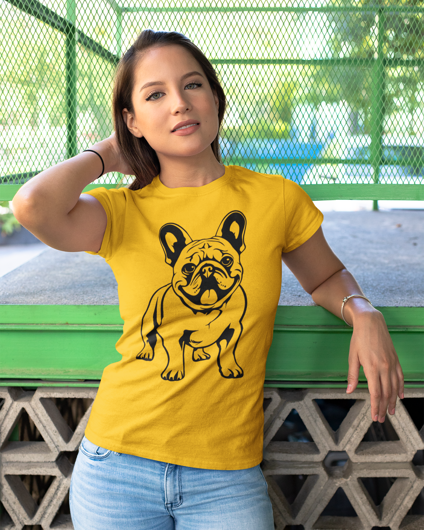 Woman wearing a yellow t-shirt with a black dog graphic, standing against a green metal fence.